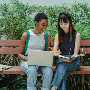 Two people sit on a park bench with notebooks to the left of them; the person on the left has a laptop in their lap, the person on the right is holding a notebook and pencil; behind them is an assortment of green foliage