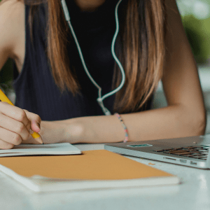 A person, head cut off by the image ending at the top, sits at a table outside with a laptop on it; they're wearing earbuds and holding a pencil over some paper and a notebook