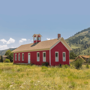 A red schoolhouse sits in the center of a green grassy field, behind it is a green tree-dotted hill and a slightly cloudy blue sky