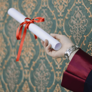 A hand holds a diploma tied with red ribbon in front of a blue and yellow patterned wall