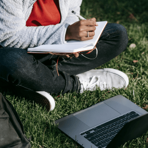 A person holding a notebook and pen sits to the left with their head cut off by the image ending; below them is a black bag and a laptop, both in the grass