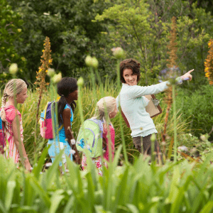 Four young children follow an adult pointing in front of them through a grassy field