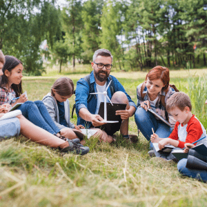 Two adults sit in grass, one holding a miniature wind turbine, surrounded by children and all in a grassy field with trees further in the background