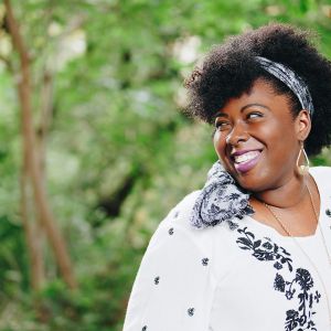A dark-skinned person in white-and-black attire looks to the left and smiles. Behind them, blurry green foliage.