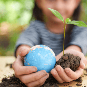 Two hands of a child reach out over a table, in the right hand is a small globe, in the left some soil with a green sprout; behind this is the child blurred with a green background