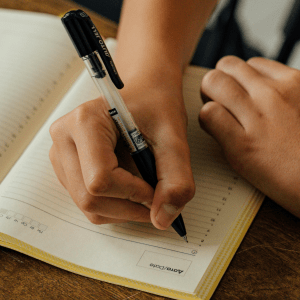 Two hands are over a notebook on a brown desk; one hand holds a pen ready to write, the other the bottom corner of the notebook