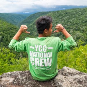 A young person sitting on the edge of a boulder overlooking forest-covered hills. The person faces away from the camera, while pointing at their green shirt that says, "YCC National Crew."