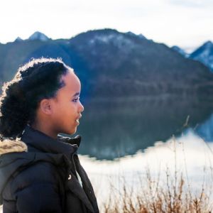 Profile of a Black child in front of a mountain lake on a partly cloudy day