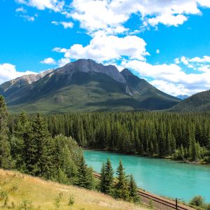 A train track alongside a river near a forest with a mountain in the background