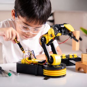 A child in a white shirt wearing safety glasses holds a screwdriver in their right hand; in front of them on a table is a yellow robotic arm