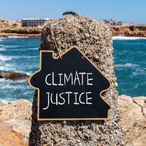 A house-shaped chalkboard with the words "Climate Justice" on it hangs from a pole in front of a sandy beach and ocean