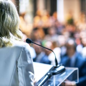 The back of a speaker looks out above a podium to a blurred crowd