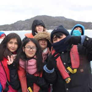 six youth bundled in warm clothing stand before a body of water with a mountain in background