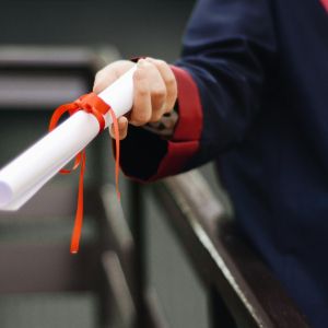 A hand holds out a rolled piece of paper tied with an orange ribbon; the person is wearing a graduation robe