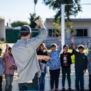 Teacher standing, hand raised, infront of a group of students in a outdoor learning space.