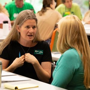 A woman in a black shirt faces another long-haired person, speaking and gesturing with her hands to herself.