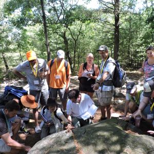 A group of people huddled in a forest with open notebooks. Some are kneeling examining a rock's surface or the ground.