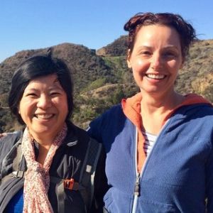 Two people stand next to each other smiling, behind them is a hilly landscape with a clear blue sky