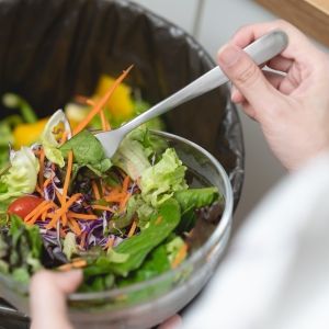 A person holds a salad bowl above a trashcan with other food waste in it.