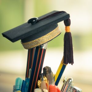 A graduate cap sits on top of pencils in a cup in front of a blurred greenish background