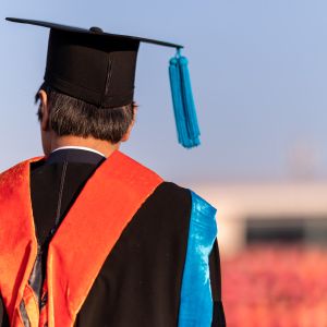 A person to the left side of the image is wearing a graduation cap and gown; to the right of them is a blurred building and background