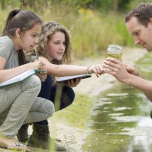 Group of three people near water. Two young women hold open notebooks while looking at a clear cup with water held by a young man.
