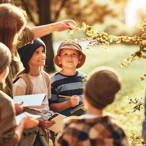 Group of curious school children with notebooks listening to their young female teacher while learning about nature together, looking at green leaf during ecology lesson in autumn forest on sunny day