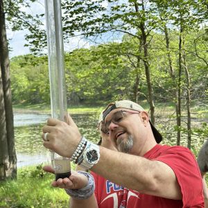 Caucasian male teacher holding a turbidity tube and smiling. There is a lake behind him surrounded by leafy trees and a blue sky.