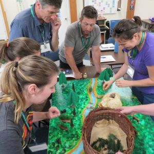 A group of five adults standing around a forested creek paper model.