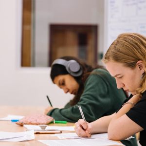 Two students work intently. One listens to something through headphones. 