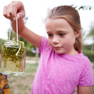 Two children look at a jar filled with water and green plants; the child on the right is holding the jar up by a string