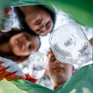 Three children look into a recycling can with a green plastic bag, putting a plastic bottle into it.