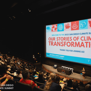 Photo of an audience sitting in an auditorium and watching a large screen of a slide that says "Our Stories of Climate Transformation"