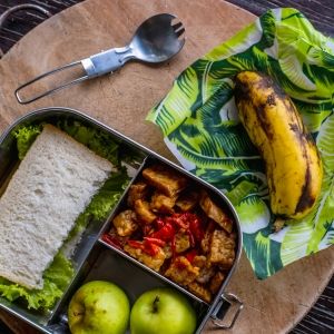 A sandwich, apples, tofu side-dish, and banana packed in an eco-friendly metal lunch box and wax paper.