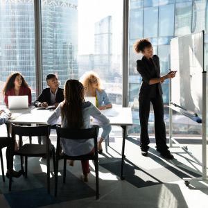 A group of 5 people sit around a rectangular table, all facing another person standing in front of a presentation board