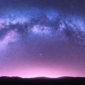 Night landscape with bright arched milky way, purple sky with stars, pink light and hills.