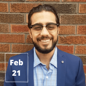 Brown-haired person smiling at the camera while in front of a brick background. On the lower left corner is a white rectangle with text inside that says, "Feb 21."