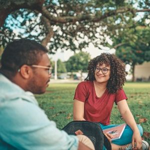 Three people sit in a grassy field talking, one with a laptop in their lap
