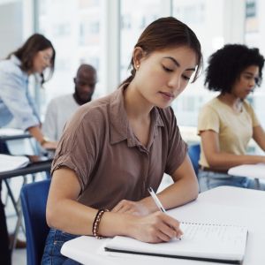 A classroom has four students sitting at a desk, with an instructor leaning over the desk of the back student