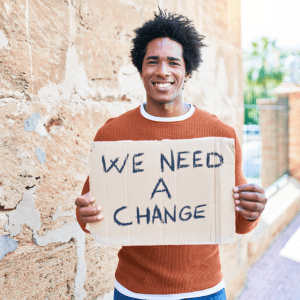 person holding "we need a change" sign on city sidewalk