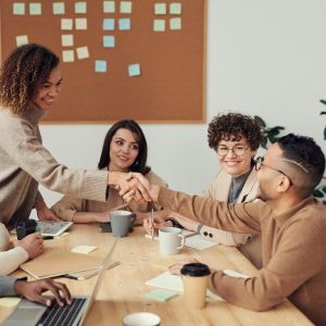 A group of six people in business casual clothes sit around a table, two reaching across the table to shake hands
