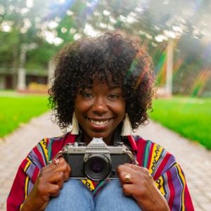 A smiling Black woman holding a camera in a park