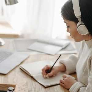 A person in front of a laptop wearing headphones and taking notes in a notebook.
