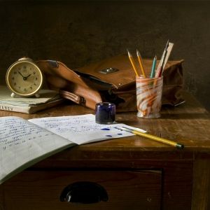 A notebook, cup full of pencils, leather bag, clock, and ink sit on a wooden desk