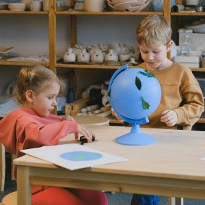 Two children interact with a globe on the table in front of them in an art classroom environment.