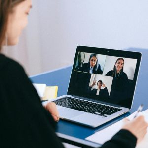 A person holding a pen over a notebook look at a laptop with a video call featuring three people
