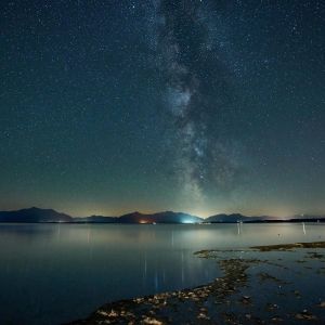 A night sky full of stars above an ocean, city lights in the distance