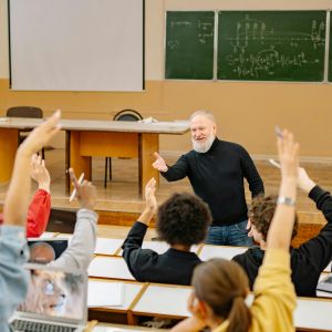 A professor standing in front of a stage with chalkboards behind it gestures to seated students raising their hands