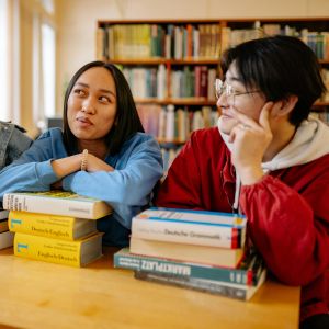 Three people sit at a table with textbooks stacked in front of them and a bookshelf behind them