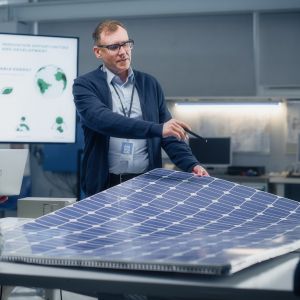 Three people are arranged around a solar panel; one person is standing and holding part of the panel with a presentation board behind them